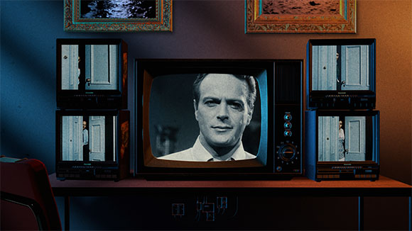 Three old-fashioned television sets on a desk; the center screen shows a man's face in black and white.