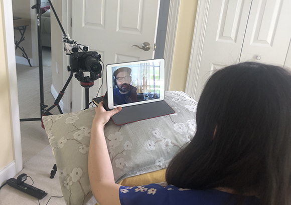A person views a man on a tablet screen propped on a pillow, with a professional camera on a tripod nearby.