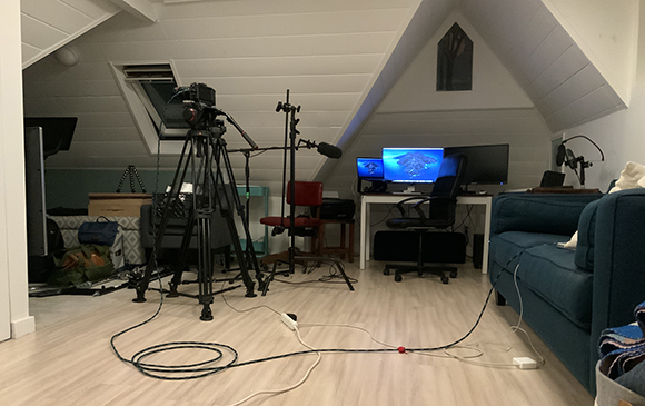 A camera on a tripod stands in a room with a computer desk, monitors, and a blue sofa.
