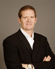 A man with light brown hair wearing a black suit and white collared shirt, posing with arms crossed.