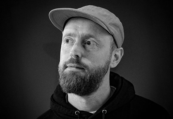 Black and white portrait of a man with a beard and a baseball cap looking upward.