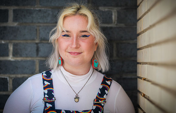A blonde woman with blue eyeliner and teal earrings smiles while wearing white turtleneck and patterned overalls.