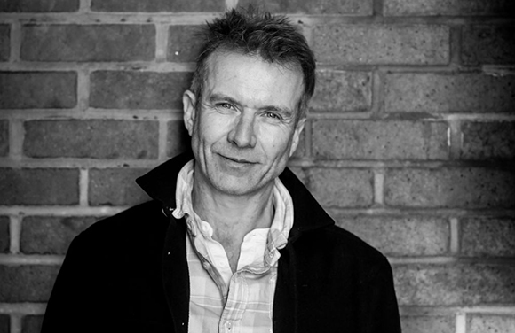 Black and white portrait of a middle-aged man smiling slightly in front of a brick wall.