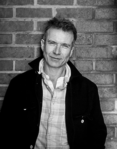 A man wearing a dark jacket over a light plaid shirt stands before a brick wall.
