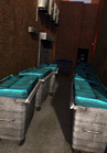 Several large, metal dumpsters with teal lids lined up in a narrow, dark alleyway.