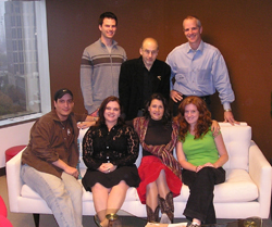 Six people, three standing and three sitting on a white sofa, posing in an office.