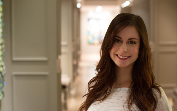 A smiling woman with long wavy brown hair wearing a white top, standing in a brightly lit hallway.