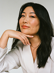 A woman with long dark hair rests her chin on her hand while wearing a white blazer.