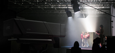 A large black digital projector in the foreground of a dark studio with stage lighting and actors in the background.