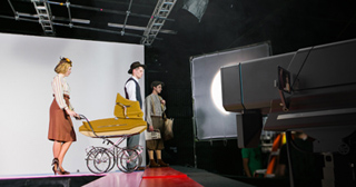 Actors in period clothing posing with a vintage baby carriage on a film set with studio lighting.