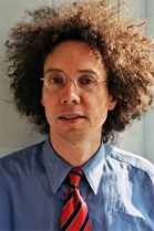 A man with voluminous curly hair, glasses, a blue shirt, and a red and navy striped tie.