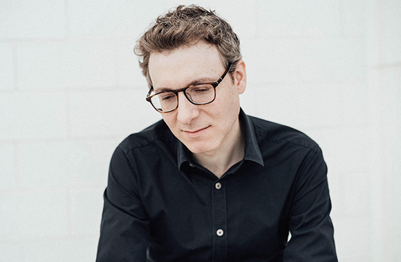 A man with wavy brown hair and glasses wearing a black button-down shirt, looking downward.