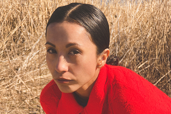 A person with dark hair pulled back wearing a bright red sweater in a field of dry, golden grass.