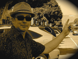 A man wearing a fedora, sunglasses, and a patterned shirt poses with one arm raised in a sepia-toned street scene.
