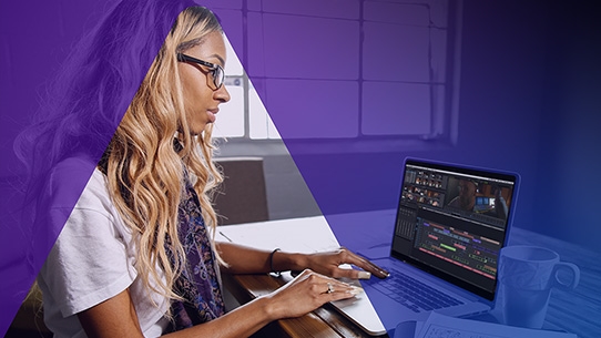 A woman with blonde hair and glasses uses a laptop to edit video in a purple-lit room.