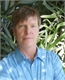 A man with short brown hair wearing a light blue button-down shirt, positioned in front of green foliage.