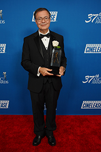 A man in a black tuxedo and glasses holds a trophy against a blue background with logos.