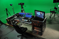 Video production studio featuring a green screen, professional camera on a tripod, and a control desk with monitors.