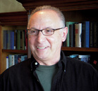 A smiling man with glasses wearing a black shirt over a green t-shirt, standing in front of a bookshelf.