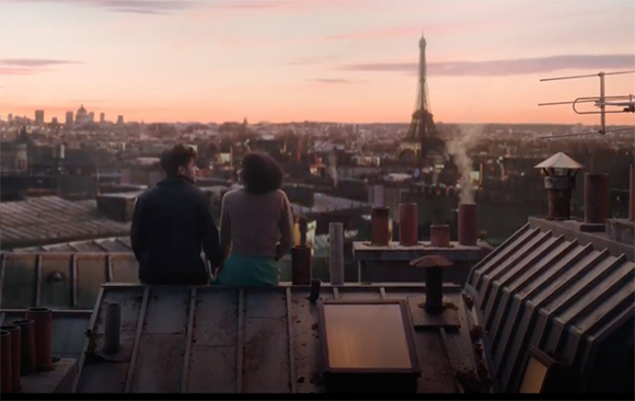 A man and woman sit on a rooftop facing the Eiffel Tower during a pink sunset over Paris.