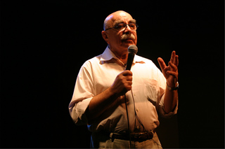 A man with glasses and a mustache speaking into a handheld microphone against a dark background.