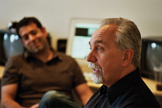 An older man with a grey goatee talks while a younger man smiles in the background.