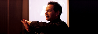 A man in a dark shirt gestures with his hands while speaking against a bright background.