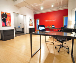 A bright office with wooden floors, a large black desk, and a red wall with text.