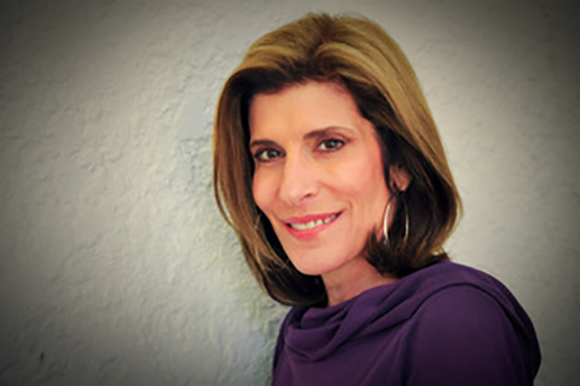 A woman with shoulder-length brown hair and silver hoop earrings smiles against a textured wall.
