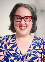 A smiling woman with short grey hair and red glasses wearing a blue and pink leaf-patterned blouse.