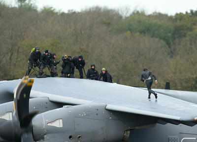 People in dark tactical gear stand and move on the wing of a large grey military aircraft.