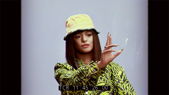 Person in a yellow bucket hat and green patterned shirt making hand gestures against a light background.