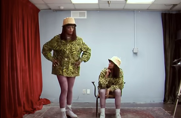 Two people wearing green animal print jackets, bucket hats, and light-colored leggings in a studio setting.