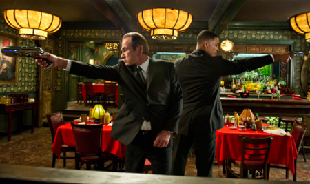 Two men in dark suits stand back-to-back, aiming handguns in a dimly lit restaurant.