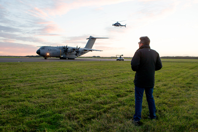 A man in a black jacket stands in a grassy field watching a large cargo plane and a helicopter.