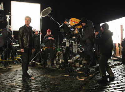 A film crew operates a large camera rig on a cobblestone street at night under bright studio lights.