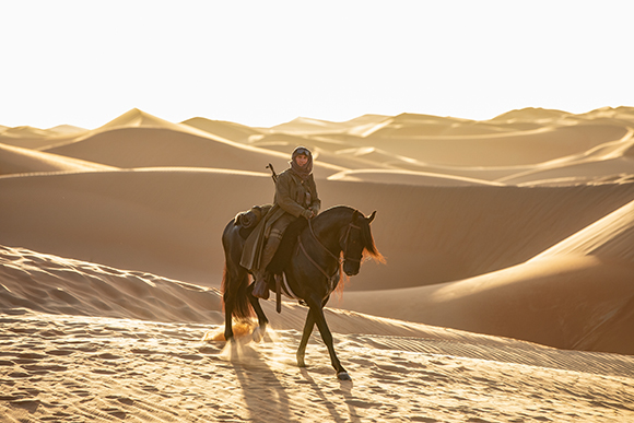 A person in a hooded cloak riding a dark horse across vast, sunlit sand dunes.