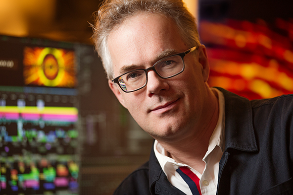 A man with short grey hair smiling in a dimly lit room with colorful digital displays in the background.
