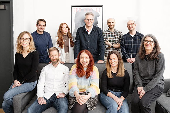 A group of eight diverse people posing together, some sitting on a couch and others standing behind them.