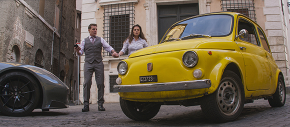 A man in a grey vest and a woman in a white shirt hold hands between a grey sports car and a yellow Fiat 500.