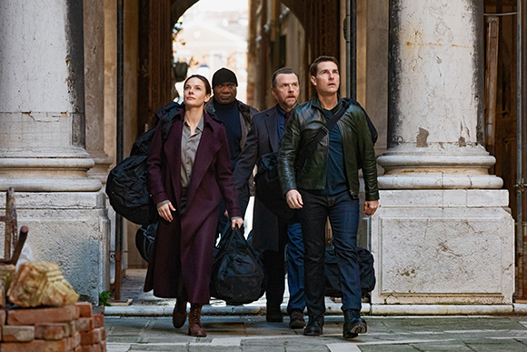 Four people walking through a stone archway carrying bags.