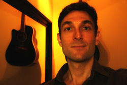 A man looking at the camera in warm, orange lighting with an acoustic guitar hanging on the wall behind him.