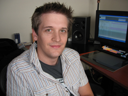 A man in a striped shirt sits in front of a computer monitor and studio speakers.
