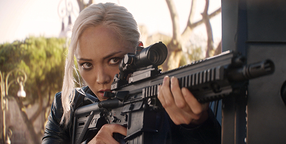 A blonde woman in a dark jacket aims a black sniper rifle with a scope toward the camera.