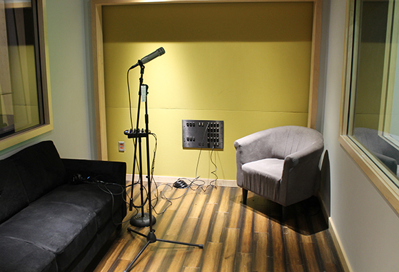 A microphone on a stand in a recording booth with a black sofa and a grey armchair.