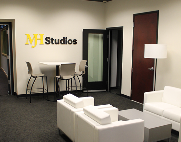 A lounge with white armchairs, a small table, and a MH Studios logo on a white wall.