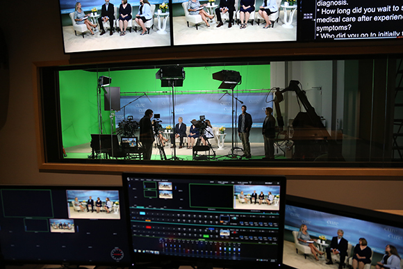 Control room monitors showing a studio set with people sitting on a couch against a green screen.
