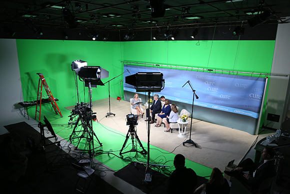 A television studio featuring a green screen, cameras, lighting, and a small group of people seated for a recording.