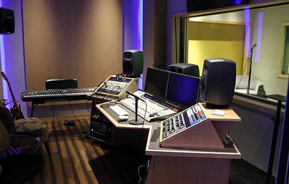 A recording studio control room with a mixing console, dual monitors, studio monitors, and musical instruments.