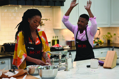 A woman prepares food in a kitchen while a man stands behind her with his hands raised in excitement.